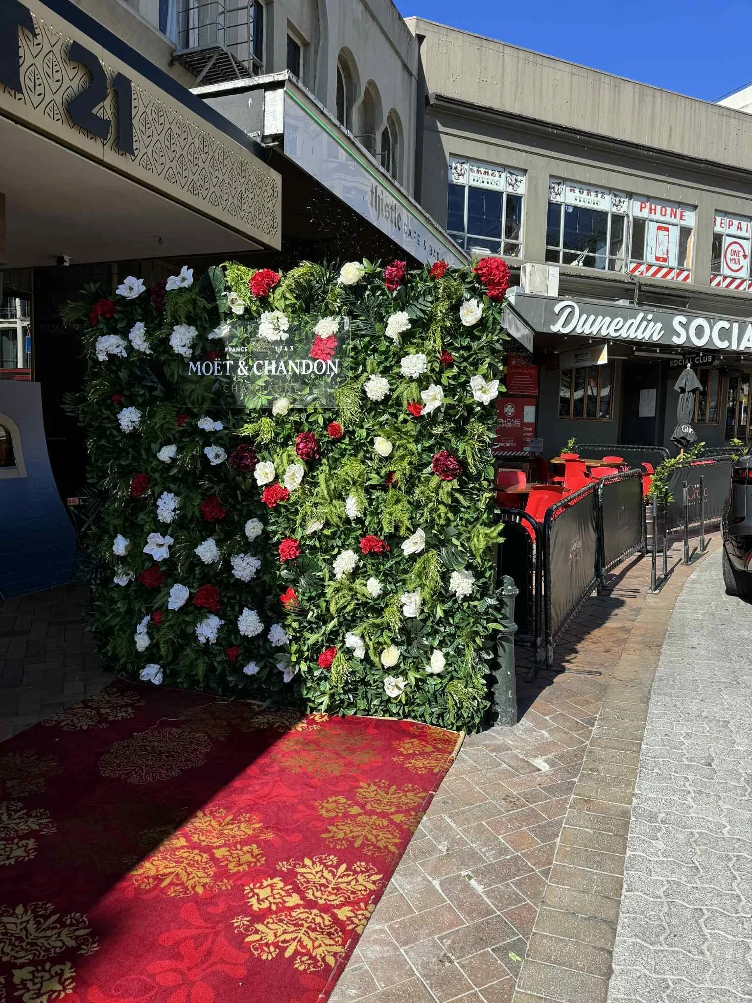 The entrance to a restaurant or bar decorated with a floral wall displaying a Moët & Chandon sign, with red and white flowers, red carpet, and outdoor seating, located on a city street with buildings and signs in the background.