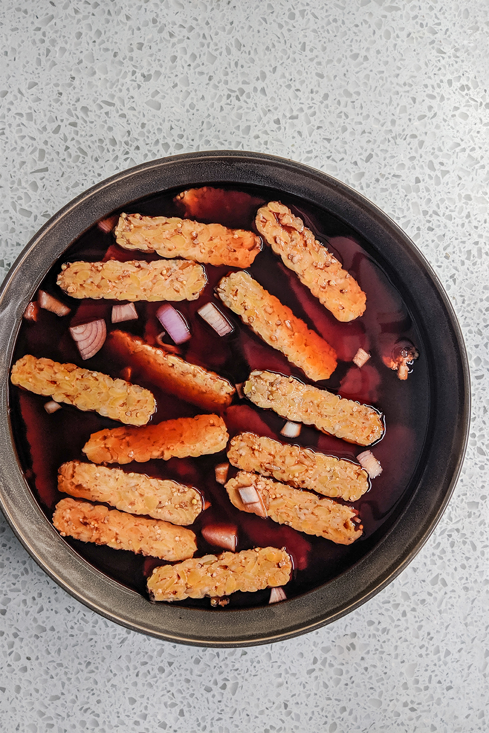  Tempeh sliced thin and marinating in beet juice, Worcestershire sauce with shallots on a grey background. 