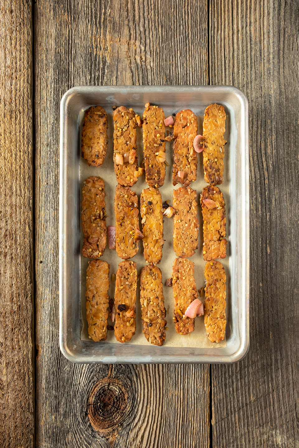 Cooked tempeh in a baking sheet lined with parchment paper on a rustic wooden background.  