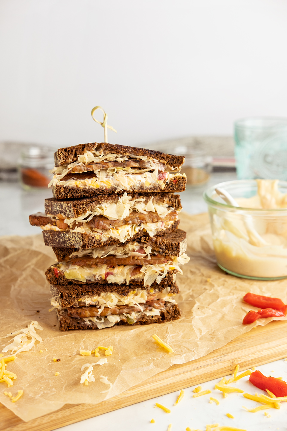  A stack of vegetarian Rueben stacked on a wooded board with parchment paper. Thousand island dressing in the background with cheese in the foreground.  