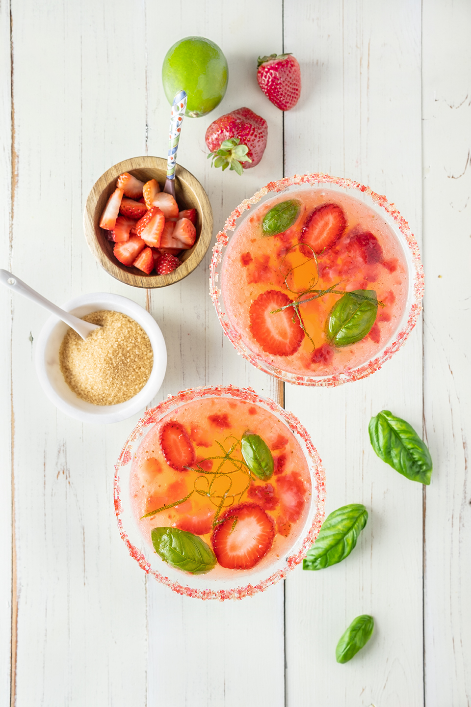  Two martini glasses filled with Strawberry Basil cocktail with sliced strawberries, basil leaves and lime zest.  Raw sugar, chopped strawberries in bowls on a rustic white wood background.  