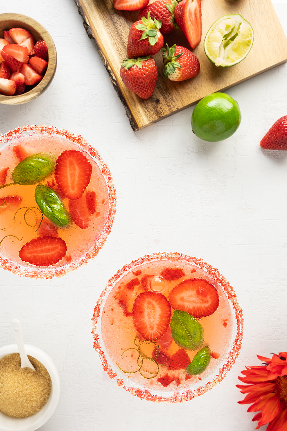  Strawberry basil cocktail in two glasses with red sugar rim.  Chopped strawberries on a cutting board with limes on a white background. 