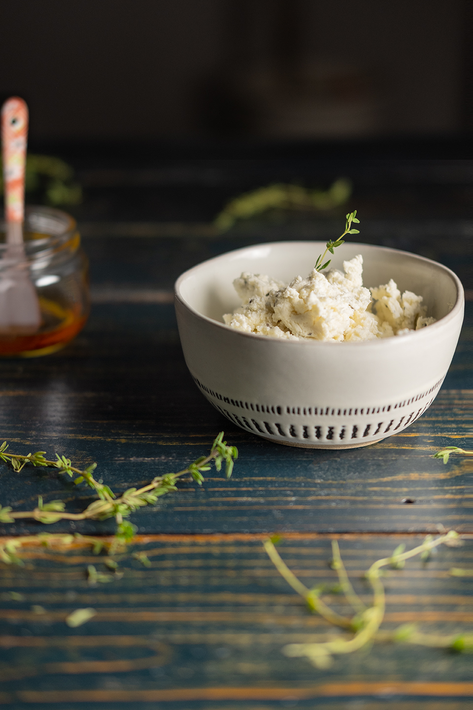  Honey,  goat cheese and thyme spread in a small bowl on a rustic blue background with honey in a jar in the background.  