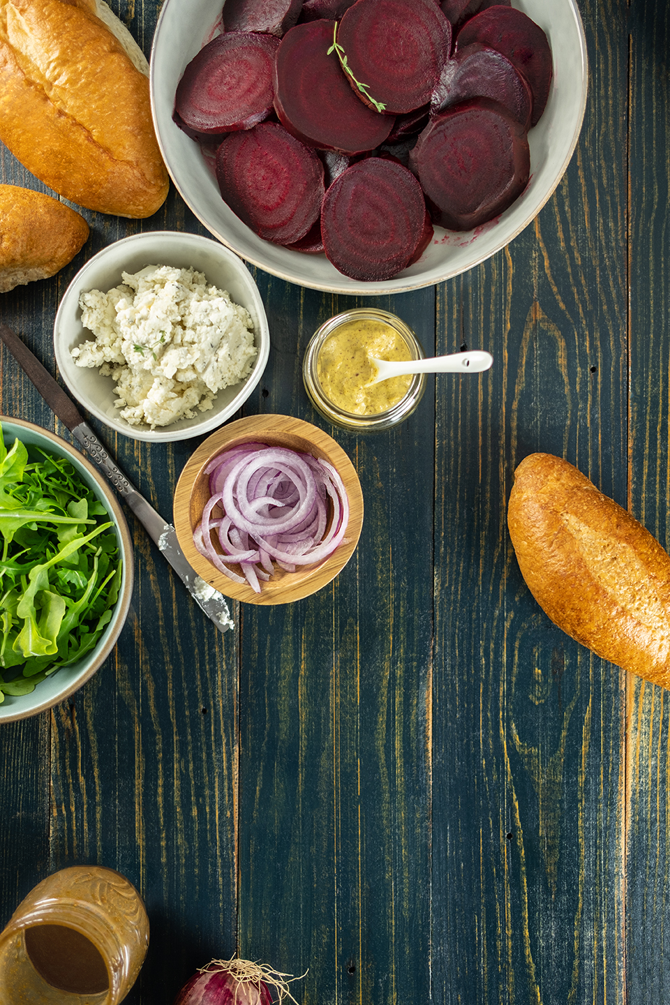  Roasted beets, honey goat cheese spread, onions, mustard, arugula and rolls on a rustic blue background.  