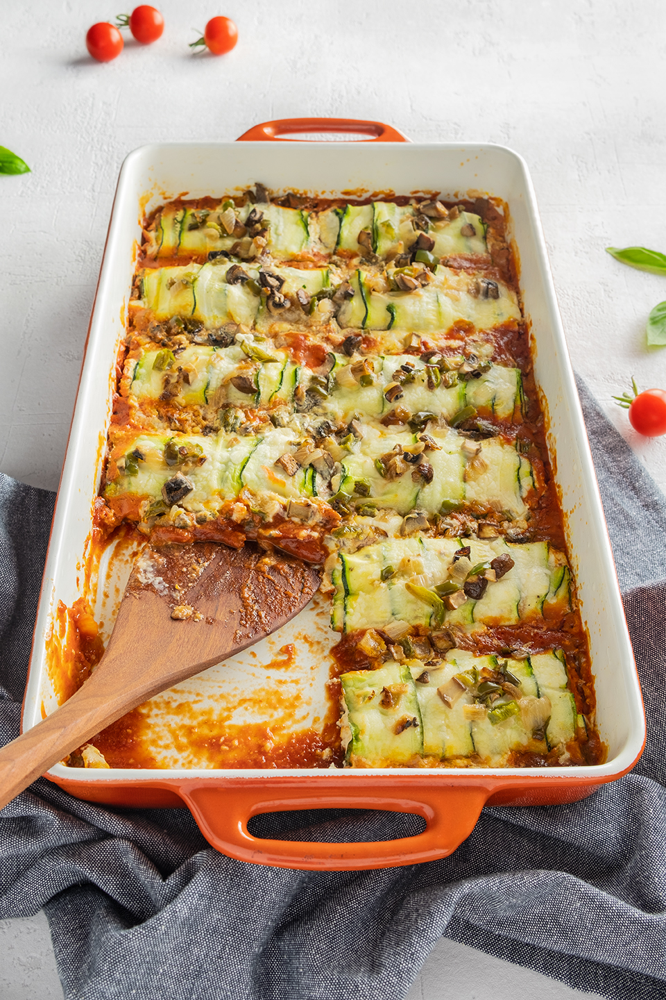  Low carb zucchini and ricotta cannelloni in a orange baking dish.  Cherry tomatoes, basil and blue napkin on a white background.  