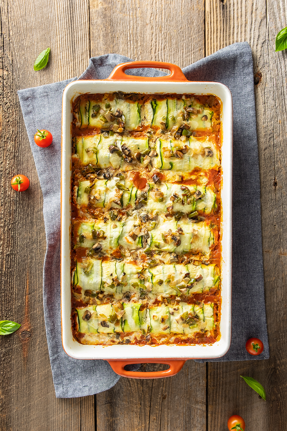  Low carb zucchini and ricotta cannelloni in a orange baking dish with a blue napkin.  Basil and cherry tomatoes on a rustic wooden background.  