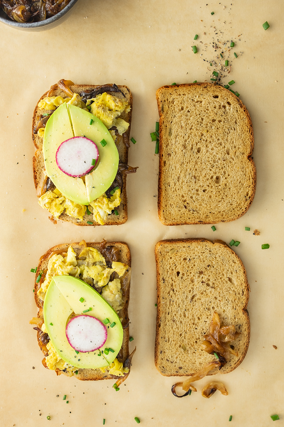  Four slices of toast, two with eggs, caramelized onions, avocado slices, radishes an chives.  Two plan pieces of toast on a brown parchment paper background.  