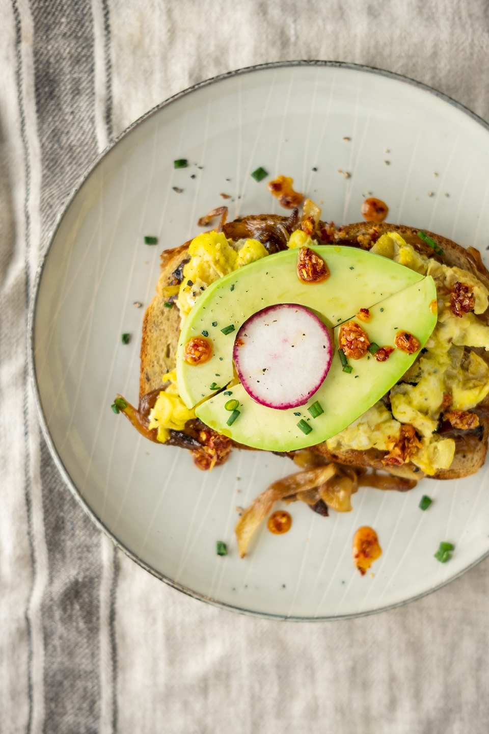  One slices of toast with eggs, caramelized onions, avocado slices, radishes, Trader Joe’s chili onion crunch and chives. Linen striped napkin under plate. 