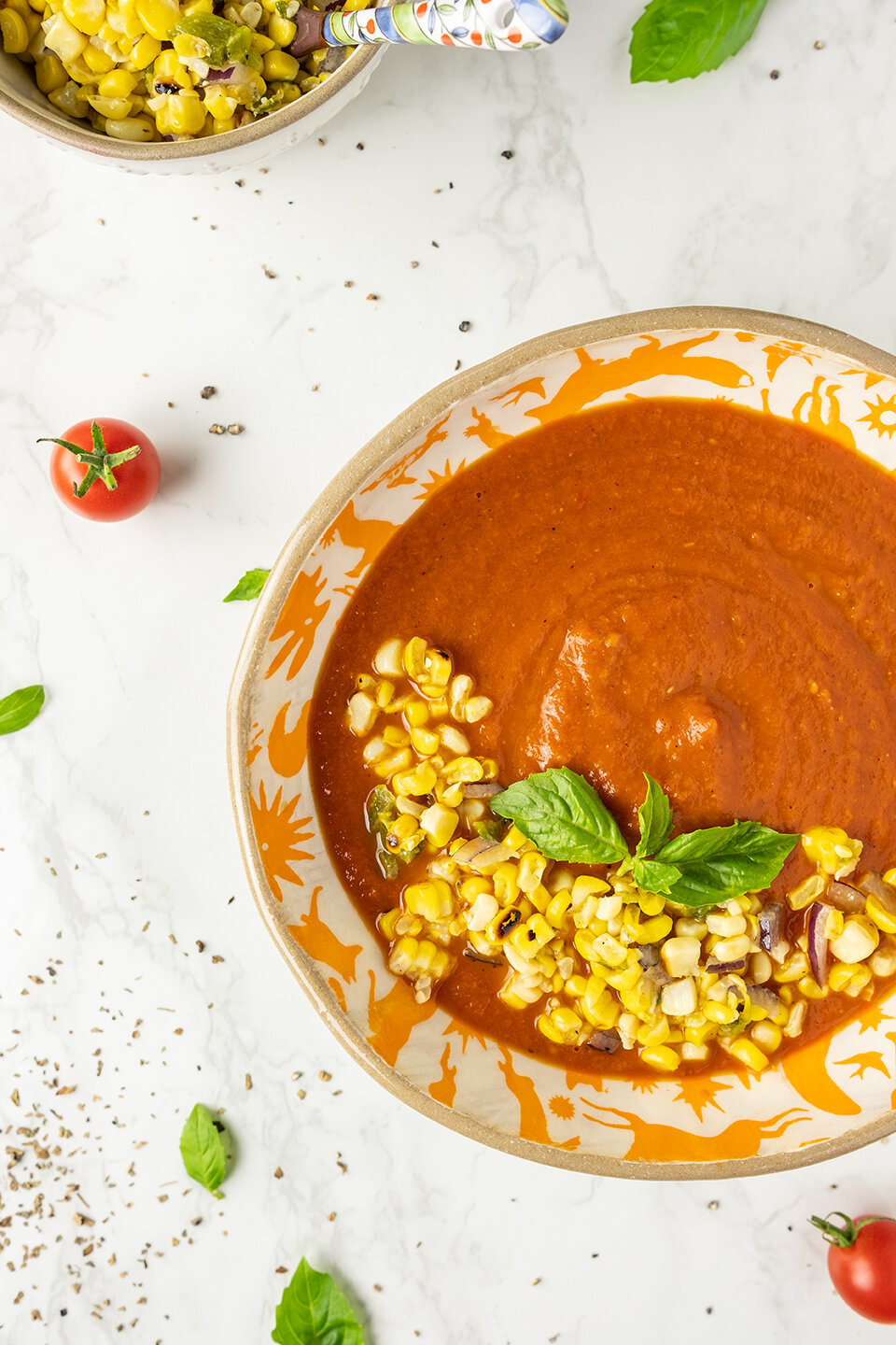  Fresh Tomato Soup with Corn Relish on a marble background. Tomatoes and basil in the background.  Bowl of corn relish with small spoon. 