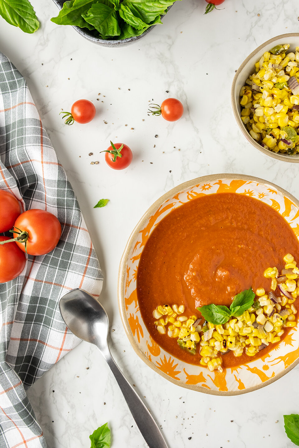  Fresh Tomato Soup with Corn Relish on a marble background.  Tomatoes and basil in the background. 