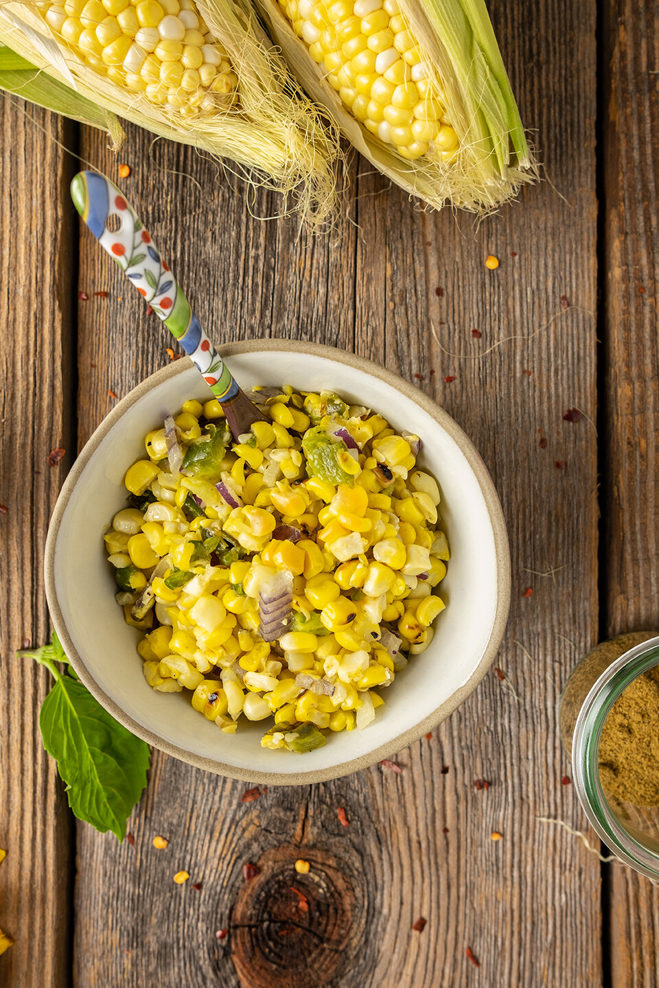  Small bowl of corn relish with colorful spoon and corn on the cob.  