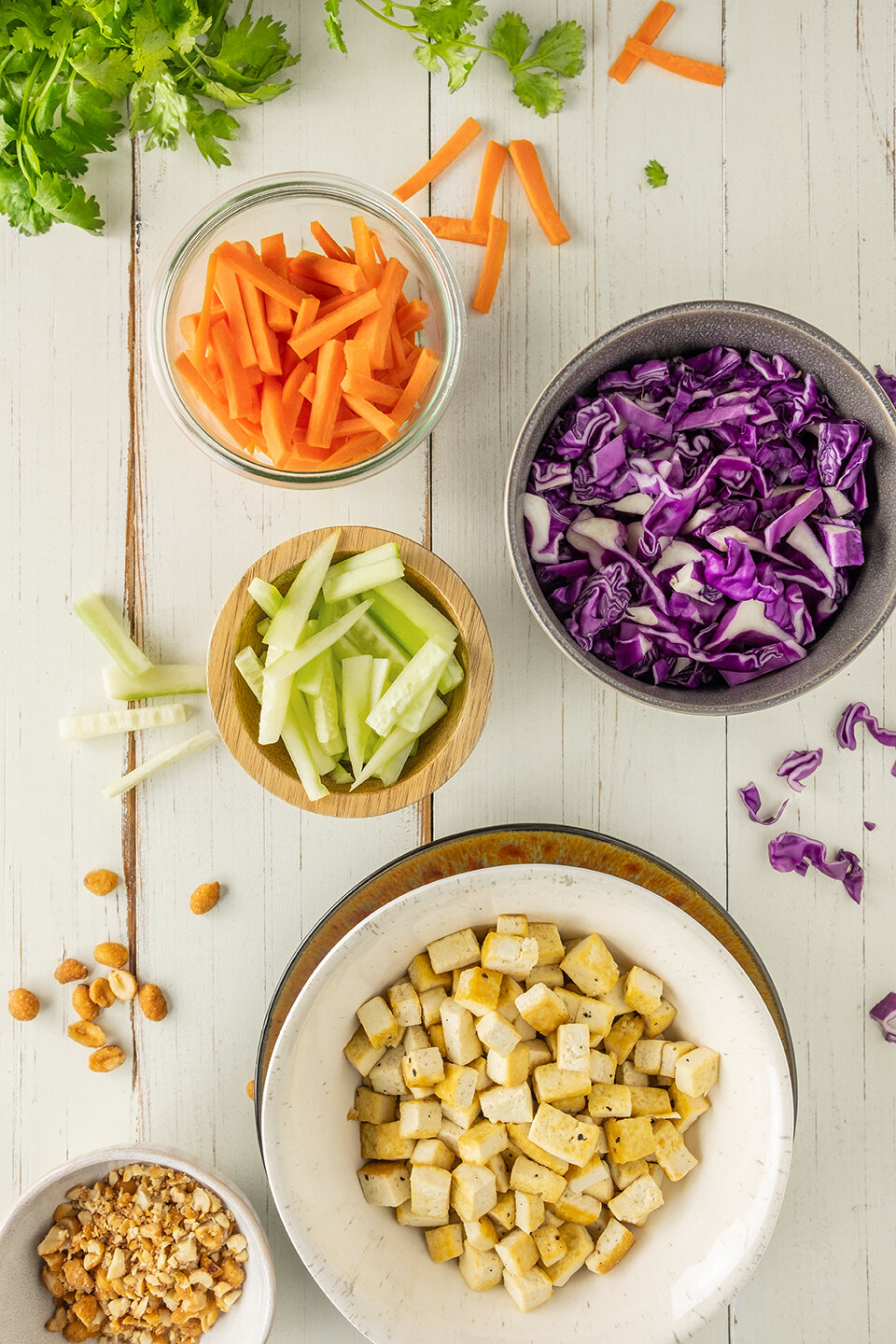  Julienned Carrots and cucumbers in small bowls.  Shredded cabbage, baked tofu cubes and cilantro on a white wooden background with a sprinkle of peanuts.  