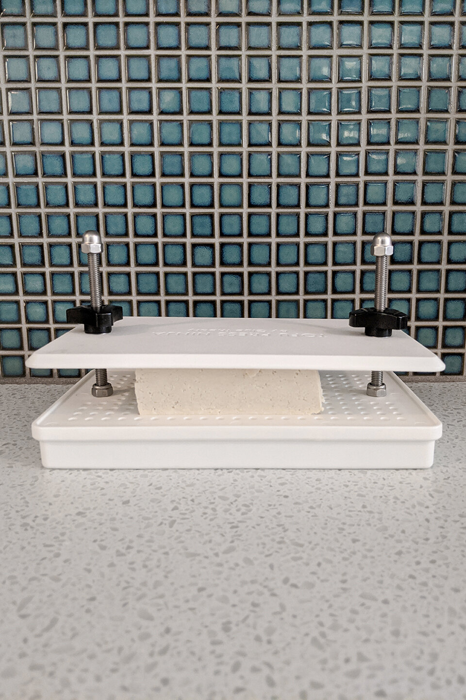  Tofu being pressed on a white counter top with blue tiled background. 