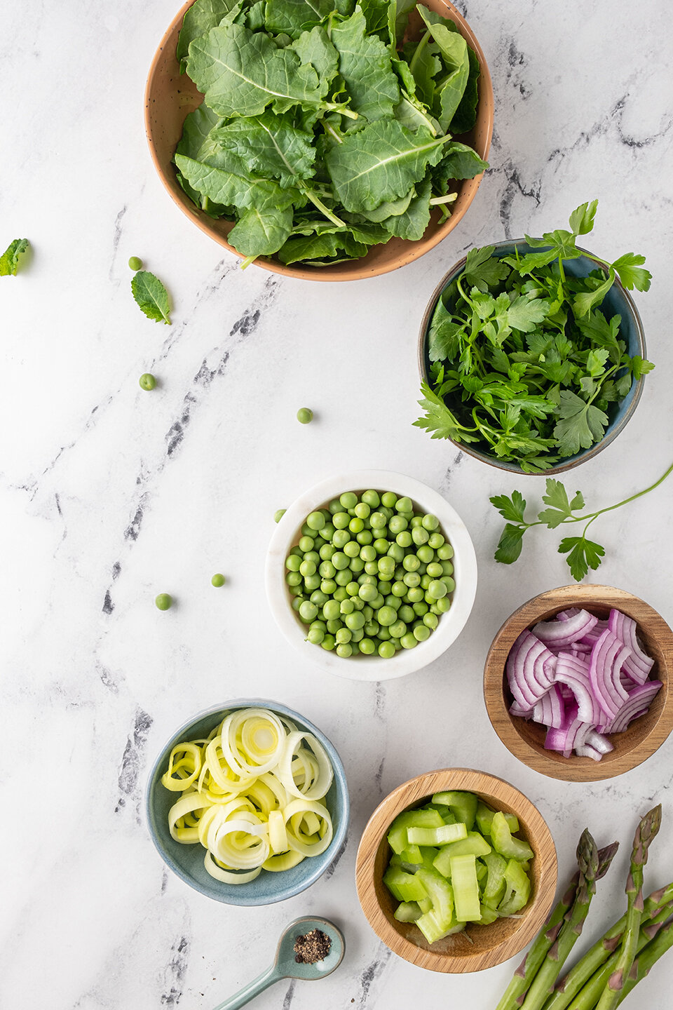  White marble background with bowls of kale, peas, parsley, red onions, leeks, celery and asparagus. 