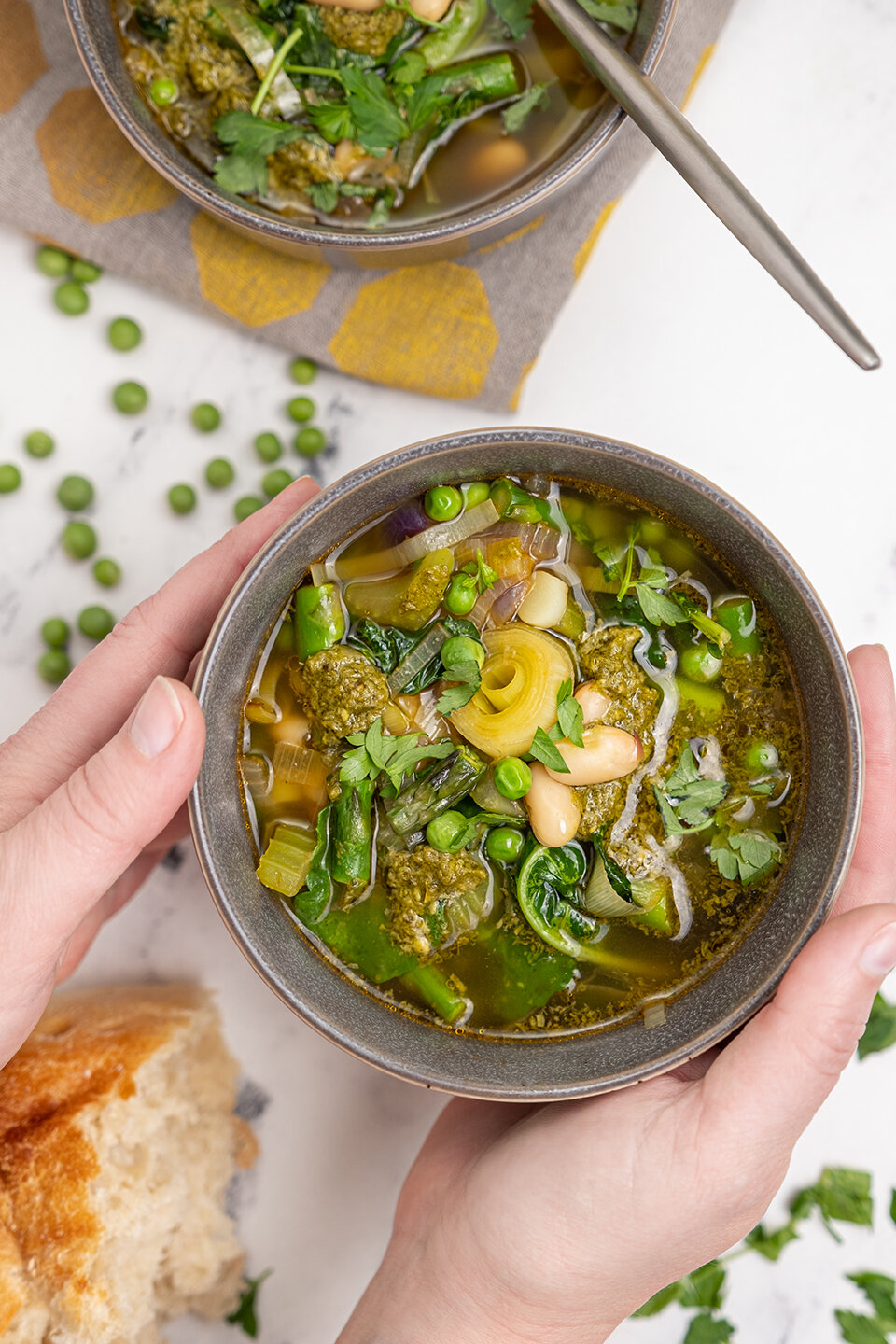  Hands holding bowl of Spring Soup with white beans.  Peas, parsley and rustic loaf of bread on a white marble background. 