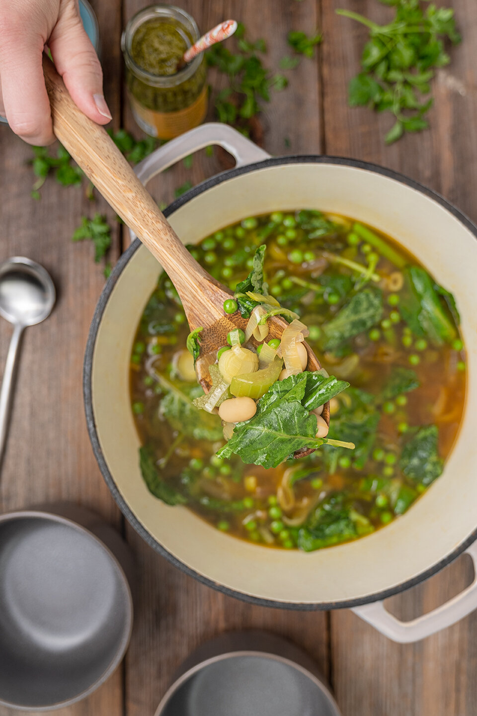  Grey dutch oven with soup and hand ladling soup into dark grey bowls. Jar of pesto and parsley on a rustic wood background.  