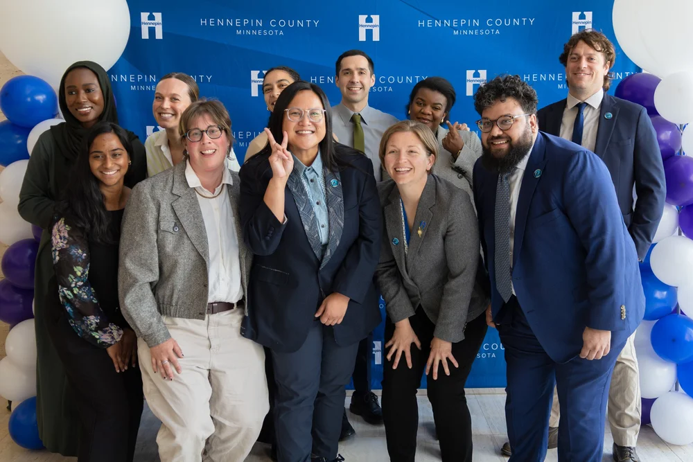 Commissioner Irene Fernando stands with a group of colleagues in front of a Hennepin County backdrop, flanked by blue, purple, and white balloons.