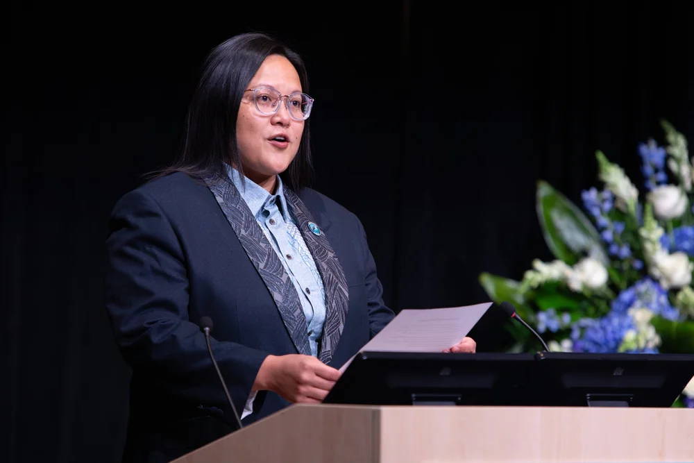 Commissioner Irene Fernando, wearing a dark blazer and a patterned scarf, speaking at a podium with floral arrangements nearby during her State of the County Address.