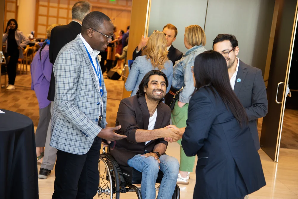 Commissioner Irene Fernando shakes hands with a seated man using a wheelchair at the 2025 State of the County address, with other attendees talking nearby.