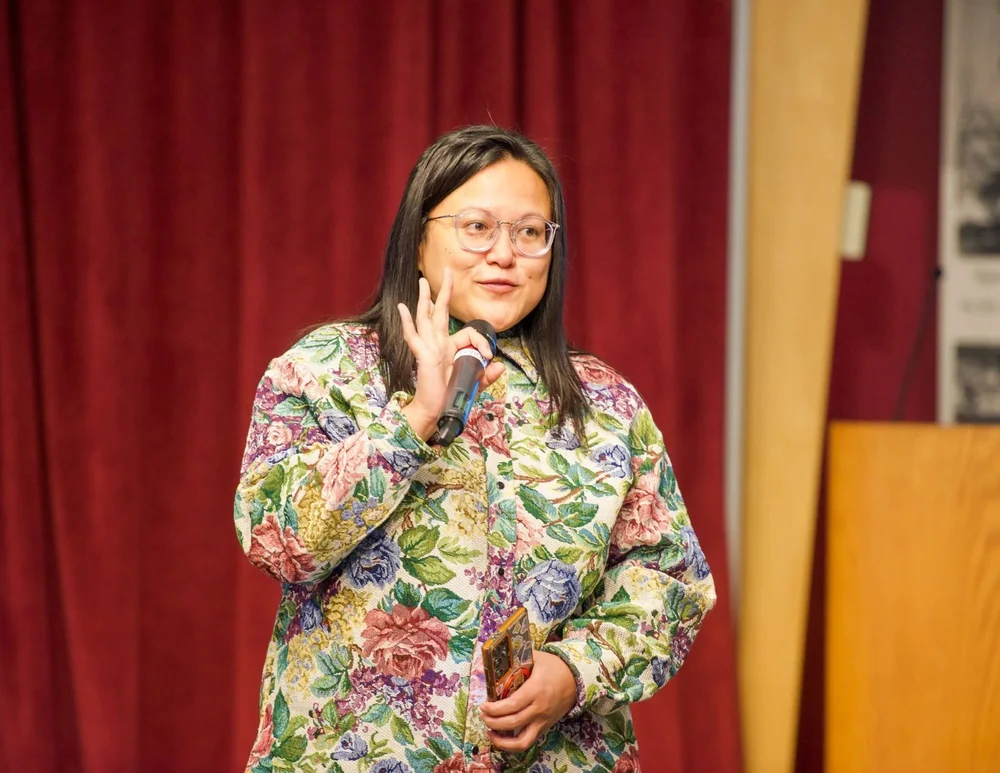 Commissioner Irene Fernando speaks into a handheld microphone, gesturing with her other hand, in front of a red stage curtain.