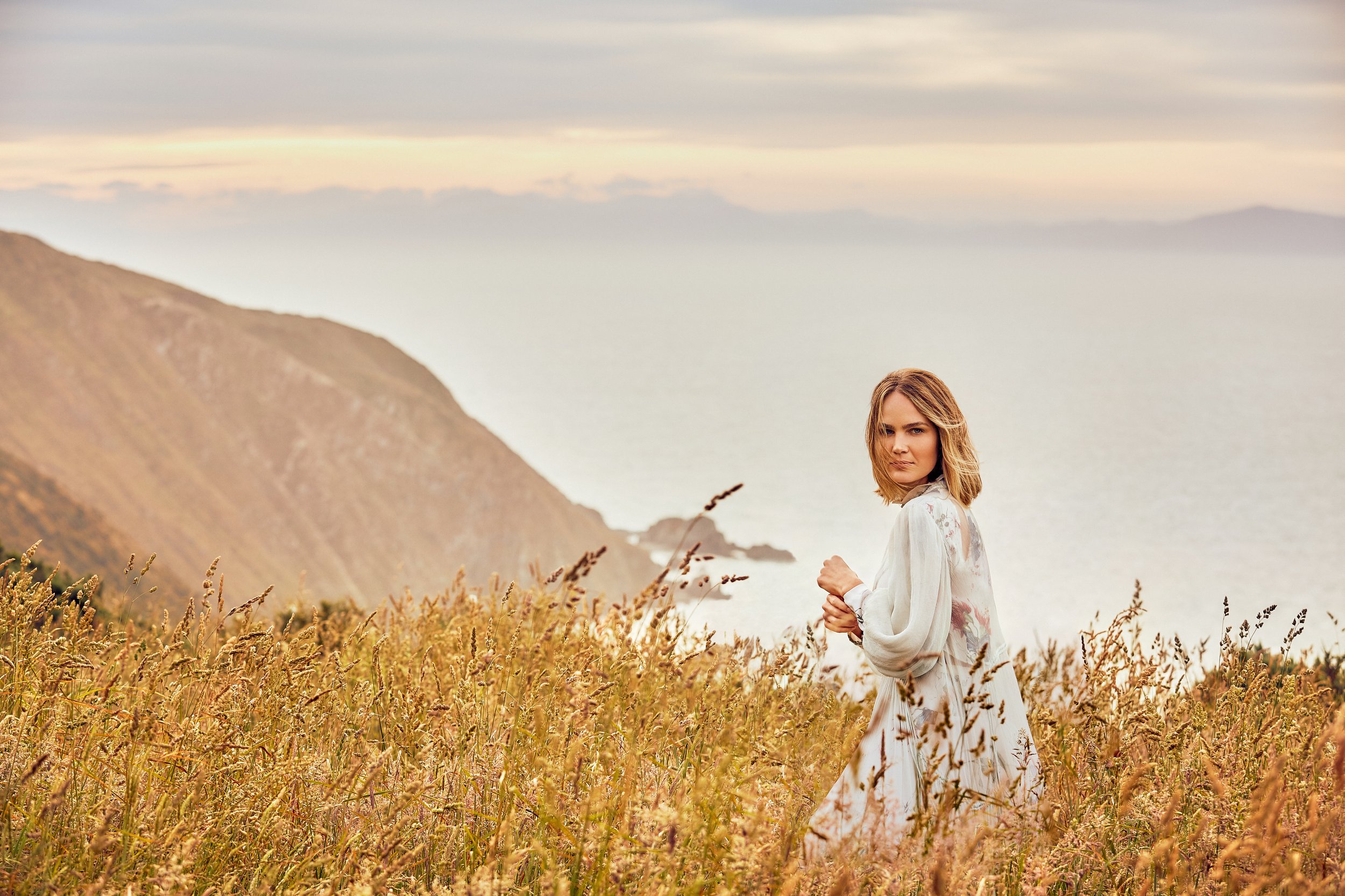 A woman with shoulder-length hair, wearing a long, flowing white dress with floral patterns, standing in a field of tall golden grass near the coast with hills and the ocean in the background under a cloudy sky.
