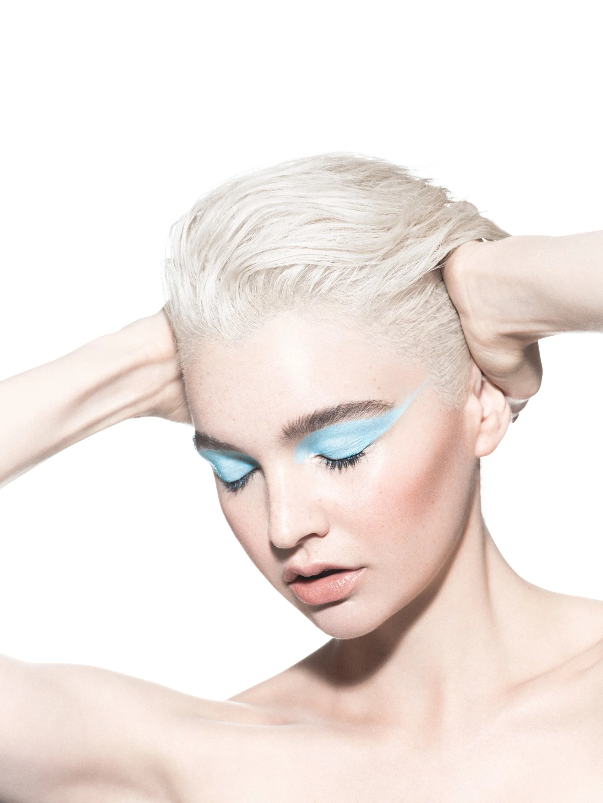 A woman with short, platinum blonde hair, her eyes closed, wearing bright blue eyeshadow, with her hands behind her head, against a white background.