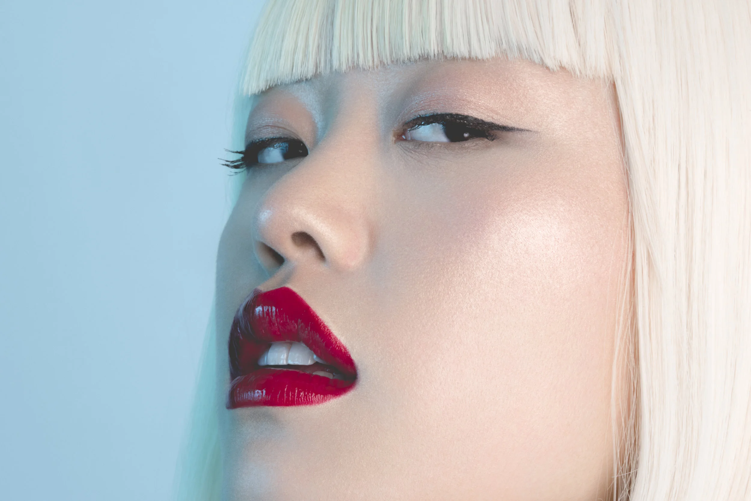 Close-up of a woman with platinum blonde hair in a blunt cut, wearing bold red lipstick, with her eyes partly closed, against a soft blue background.