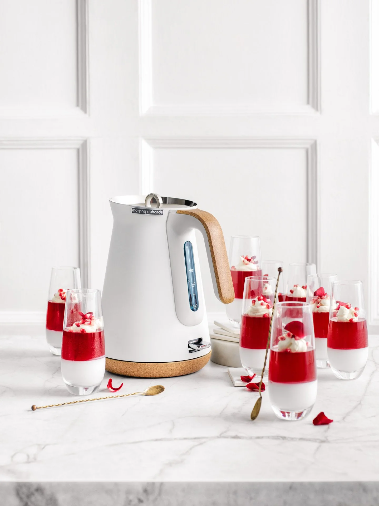 A white modern kettle with a wooden handle and a small spout, surrounded by several glasses of layered red and white desserts topped with whipped cream and small red decorations on a white marble surface.