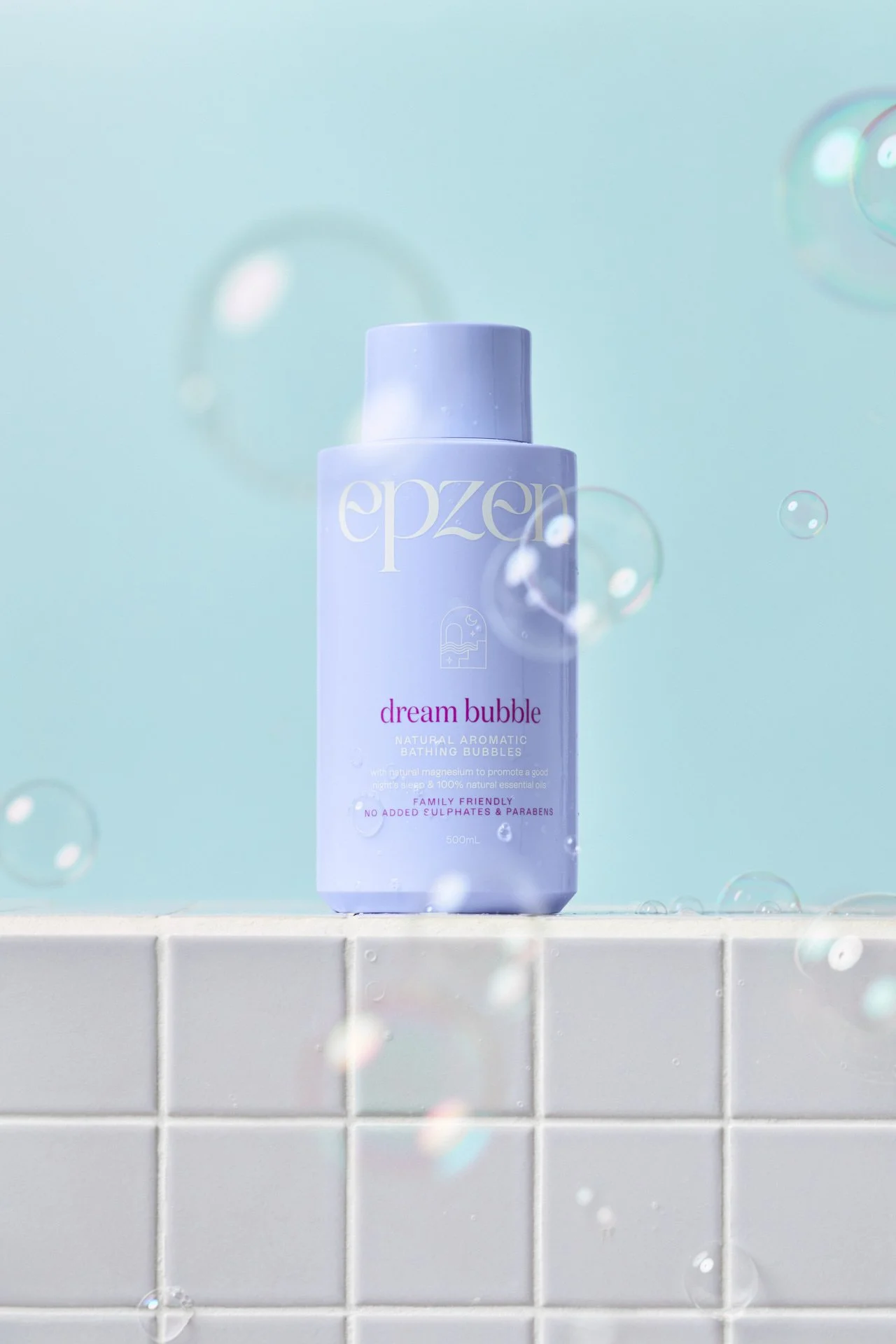A bottle of Epzen Dream Bubble natural aromatic bathing bubbles on a tiled surface with water droplets and floating bubbles, against a light blue background.