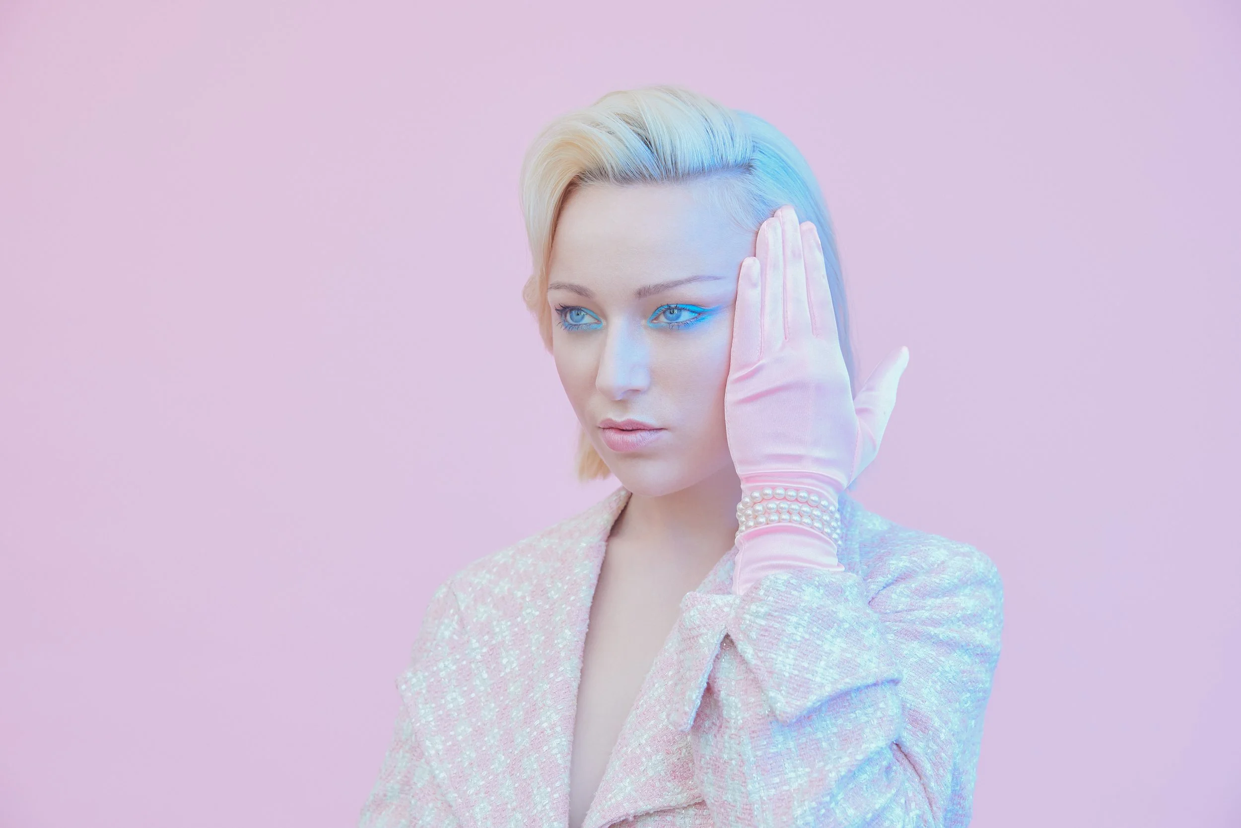 A woman with platinum blonde hair with a blue hue, wearing pink gloves, pearl bracelet, and a pastel-colored textured blazer, poses against a pastel pink background, with her hand near her face.