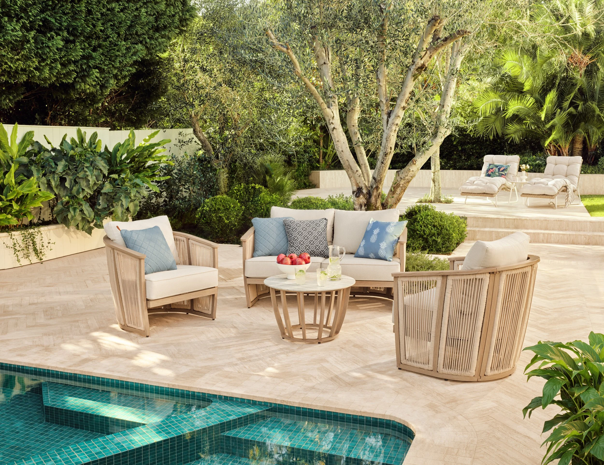 A backyard patio with outdoor seating around a swimming pool, surrounded by lush green trees and plants.