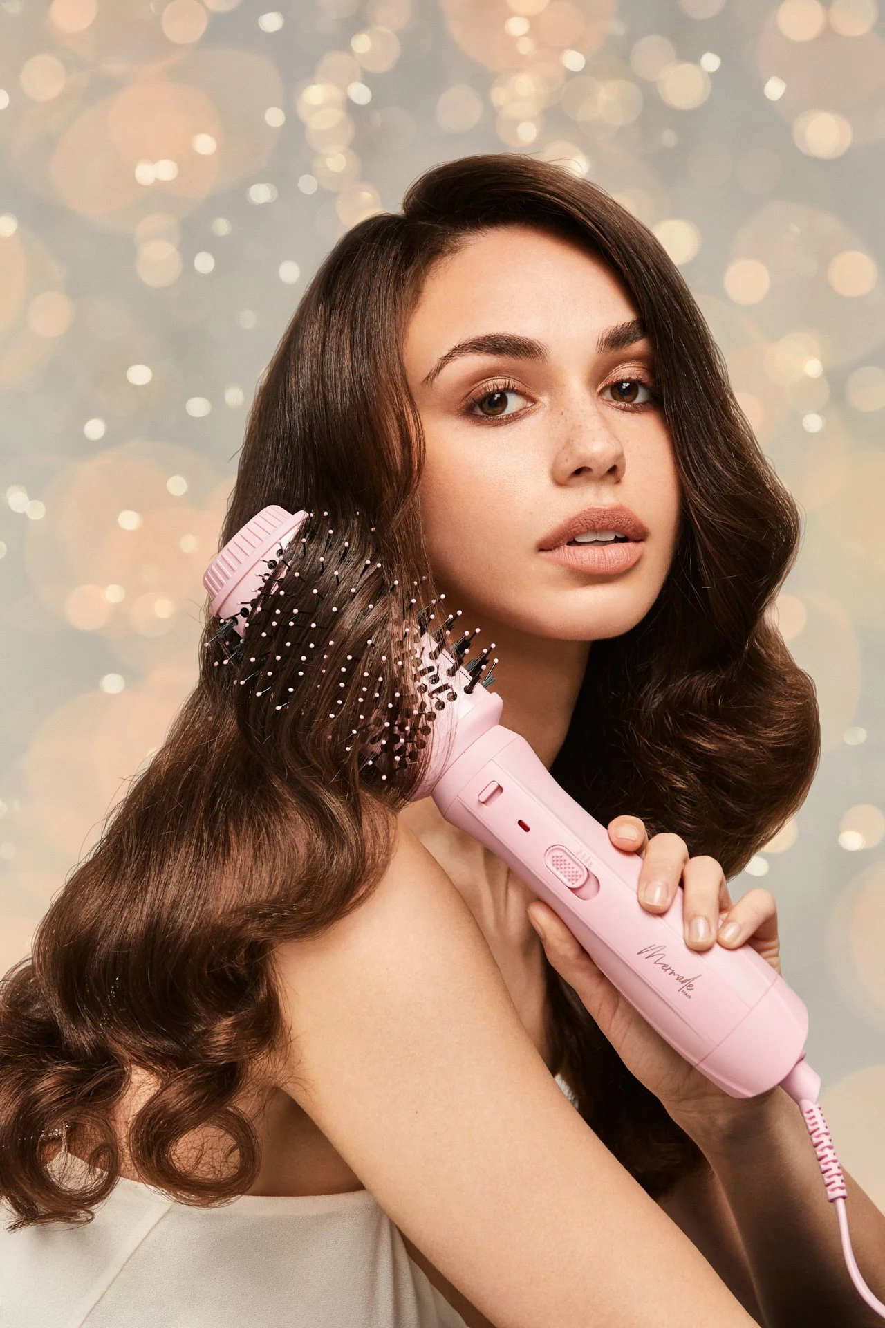 A woman with long, wavy brown hair takes a selfie while holding a pink hairbrush with a built-in hairdryer, with a bokeh background.