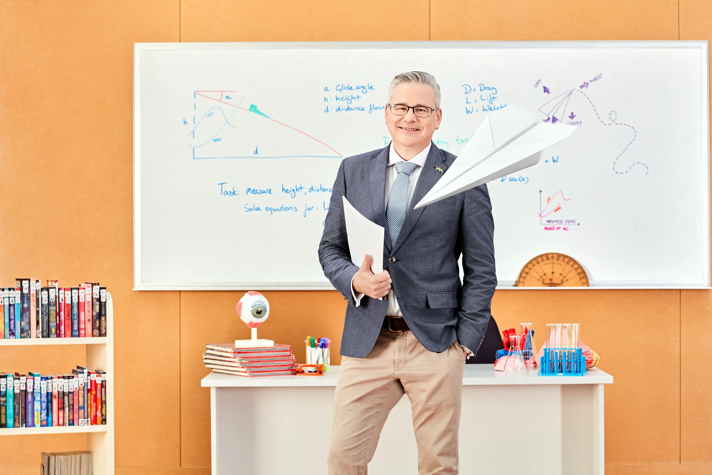 A teacher in a classroom holding papers, standing in front of a whiteboard with science and math diagrams, tables, and written notes. The classroom has shelves with books, a globe, and science lab equipment.