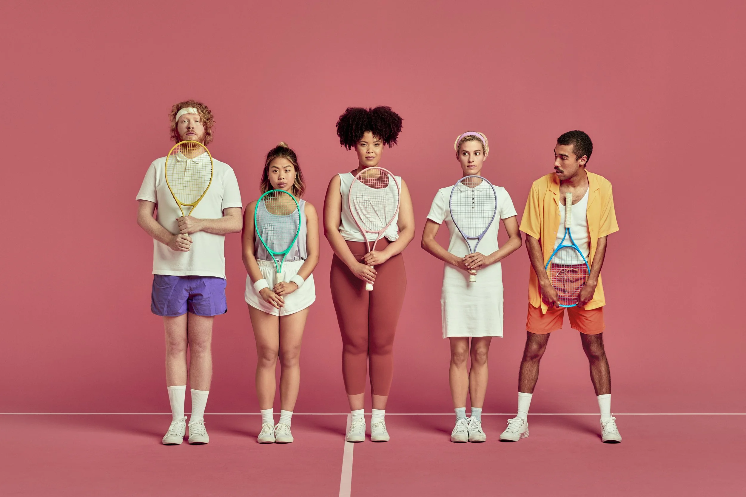 Five individuals standing on a pink badminton court, holding badminton rackets, dressed in casual sportswear, with a pink wall background.