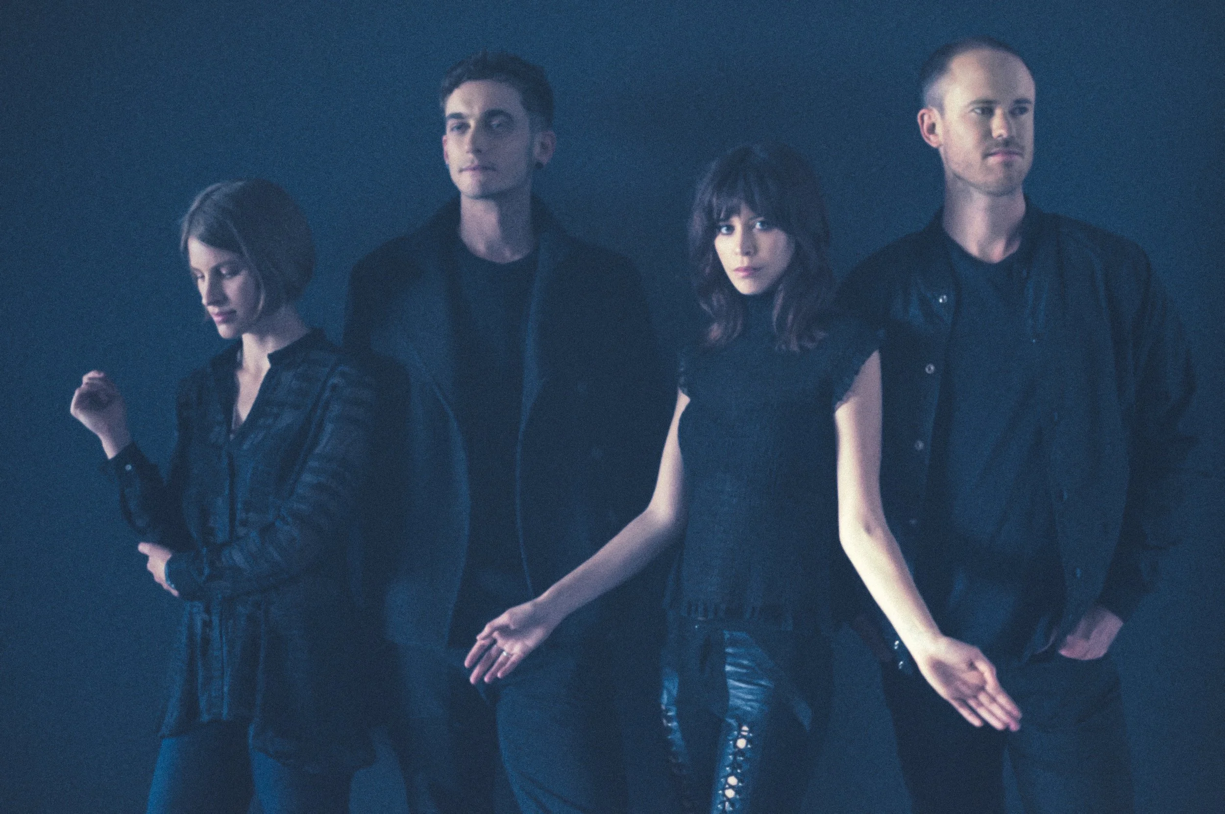 Four people standing in a line in a dark setting, wearing black clothes. The person on the far left has short hair and looks down. The second person from the left has short dark hair and is looking straight ahead. The third person has shoulder-length