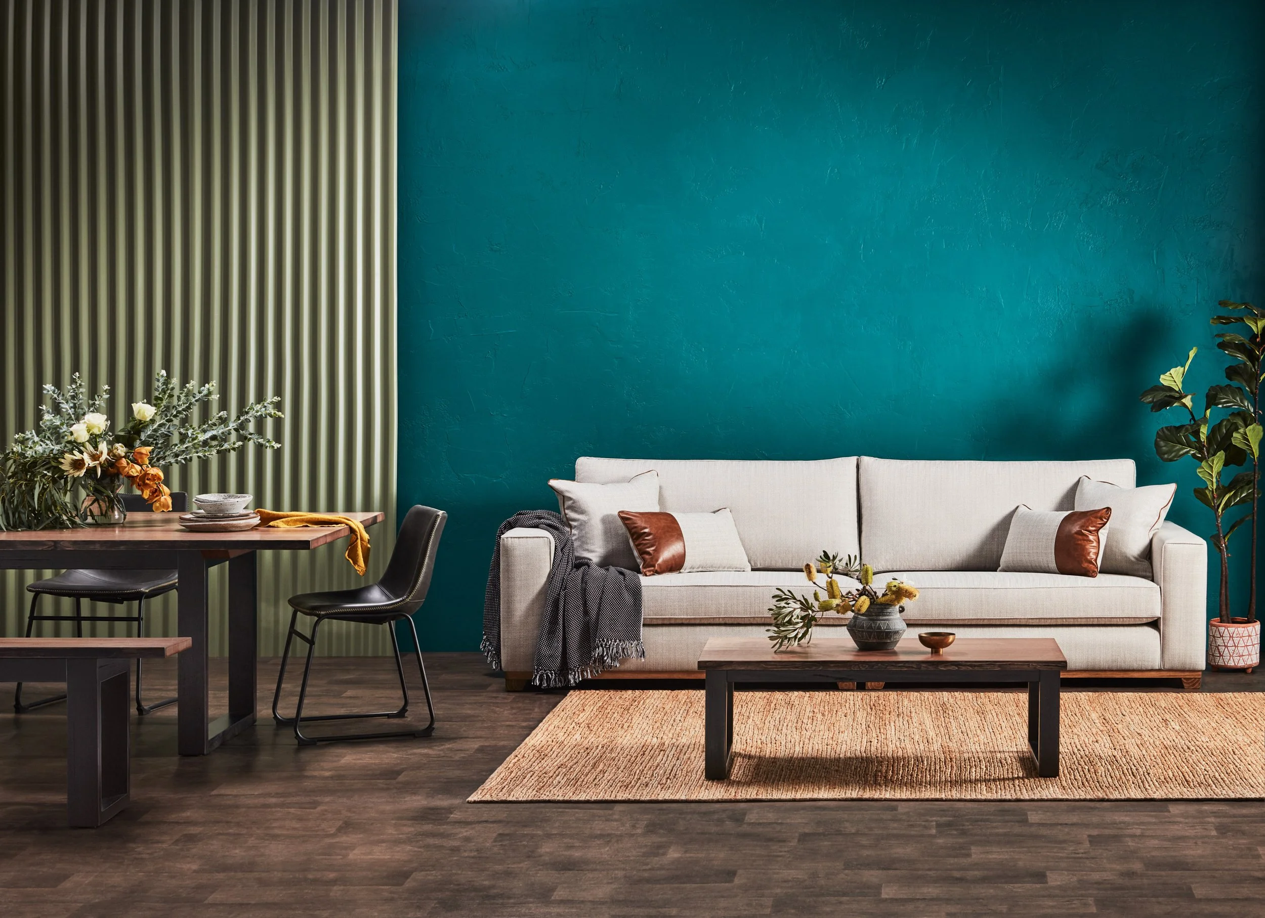Living room with a teal wall, white sofa with brown pillows, a wooden coffee table with plants, and a tall plant on the right side. A dining area with a wooden table, black chairs, and a flower arrangement is on the left side.