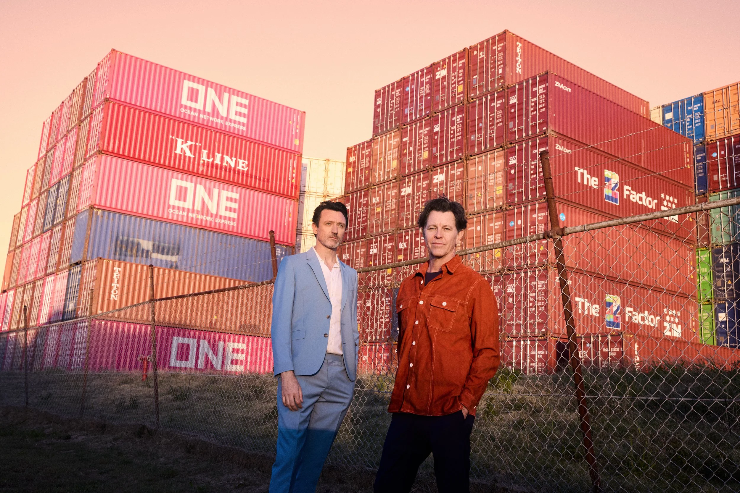 Two men stand in front of stacked cargo containers in a shipping yard during sunset. One man wears a light blue suit, and the other wears a red jacket.