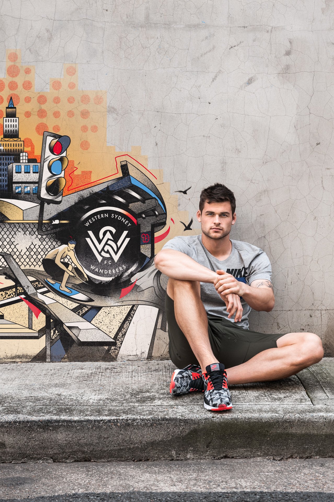 A young man sitting on the sidewalk next to a graffiti mural, wearing a gray Nike t-shirt, black shorts, and patterned sneakers, with a neutral expression.