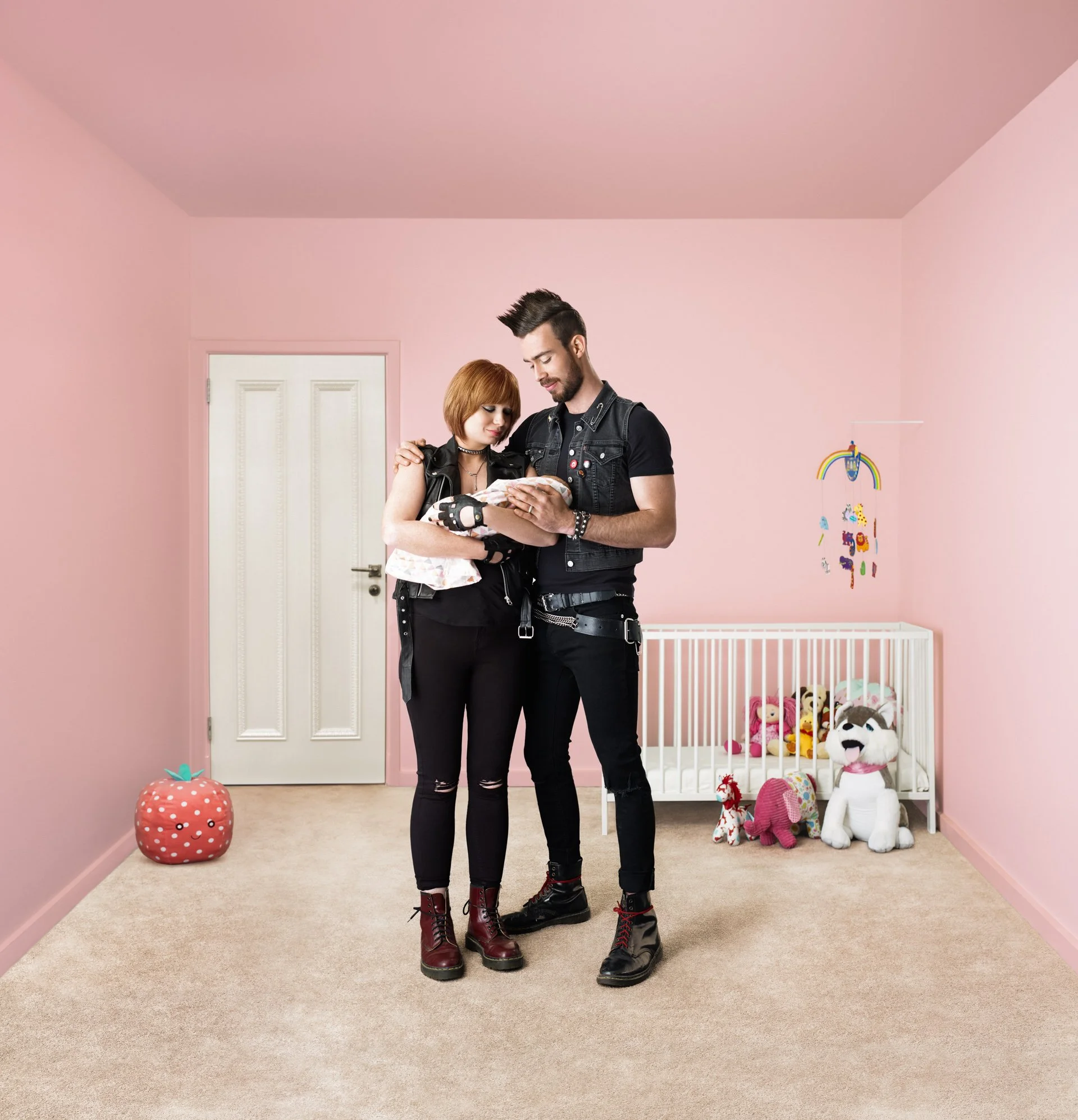 A young couple dressed in goth style standing in a pink bedroom holding a newborn baby. The woman has red hair, and both are wearing black leather outfits with combat boots. The bedroom has a white crib filled with plush toys, colorful wall decoratio