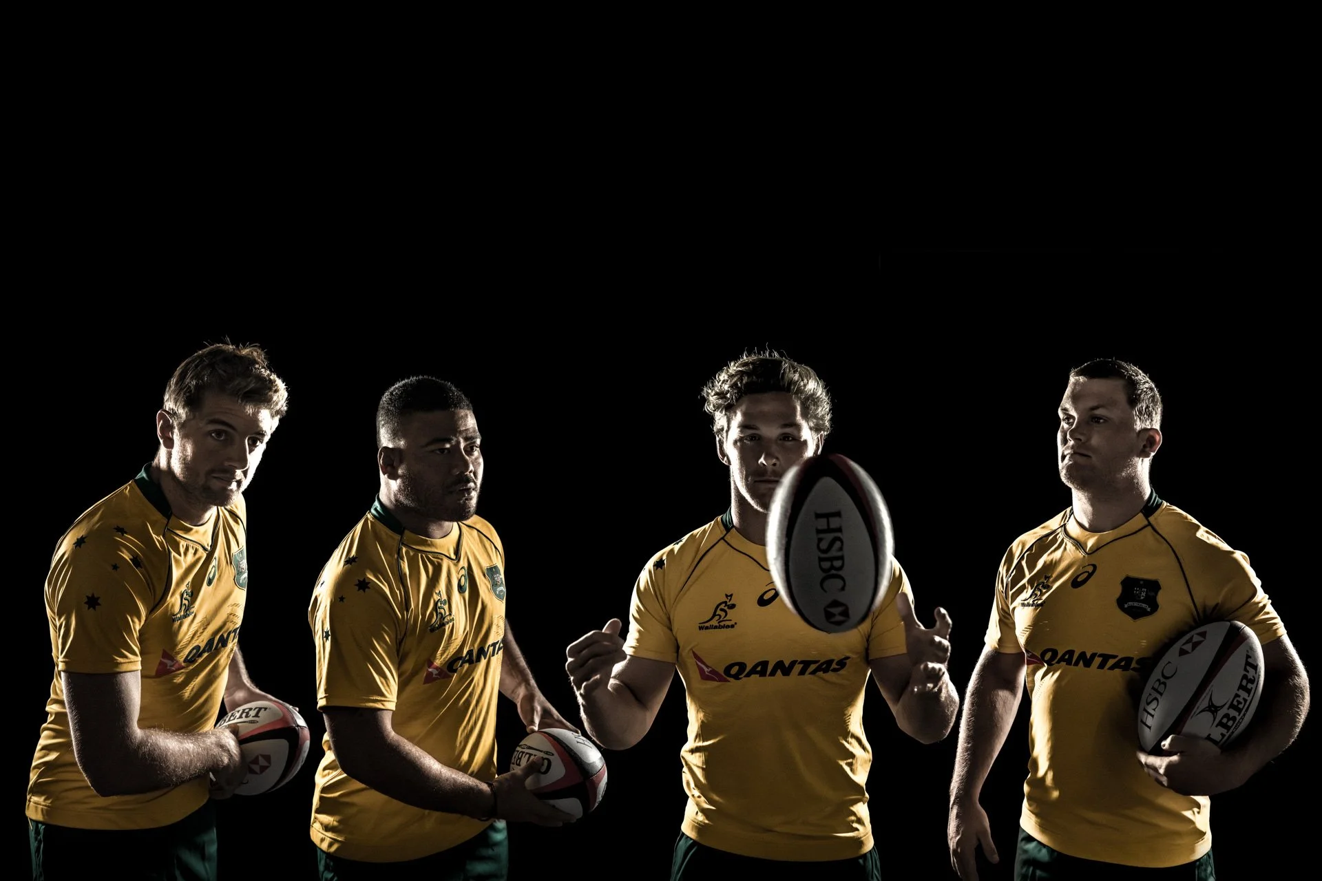 Four rugby players in yellow jerseys holding rugby balls, standing against a black background with a rugby ball in the foreground.