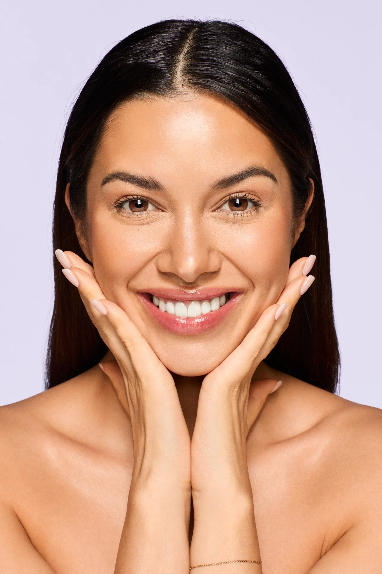 Close-up of a smiling woman with dark brown hair, brown eyes, and clear skin, holding her face gently with her hands.