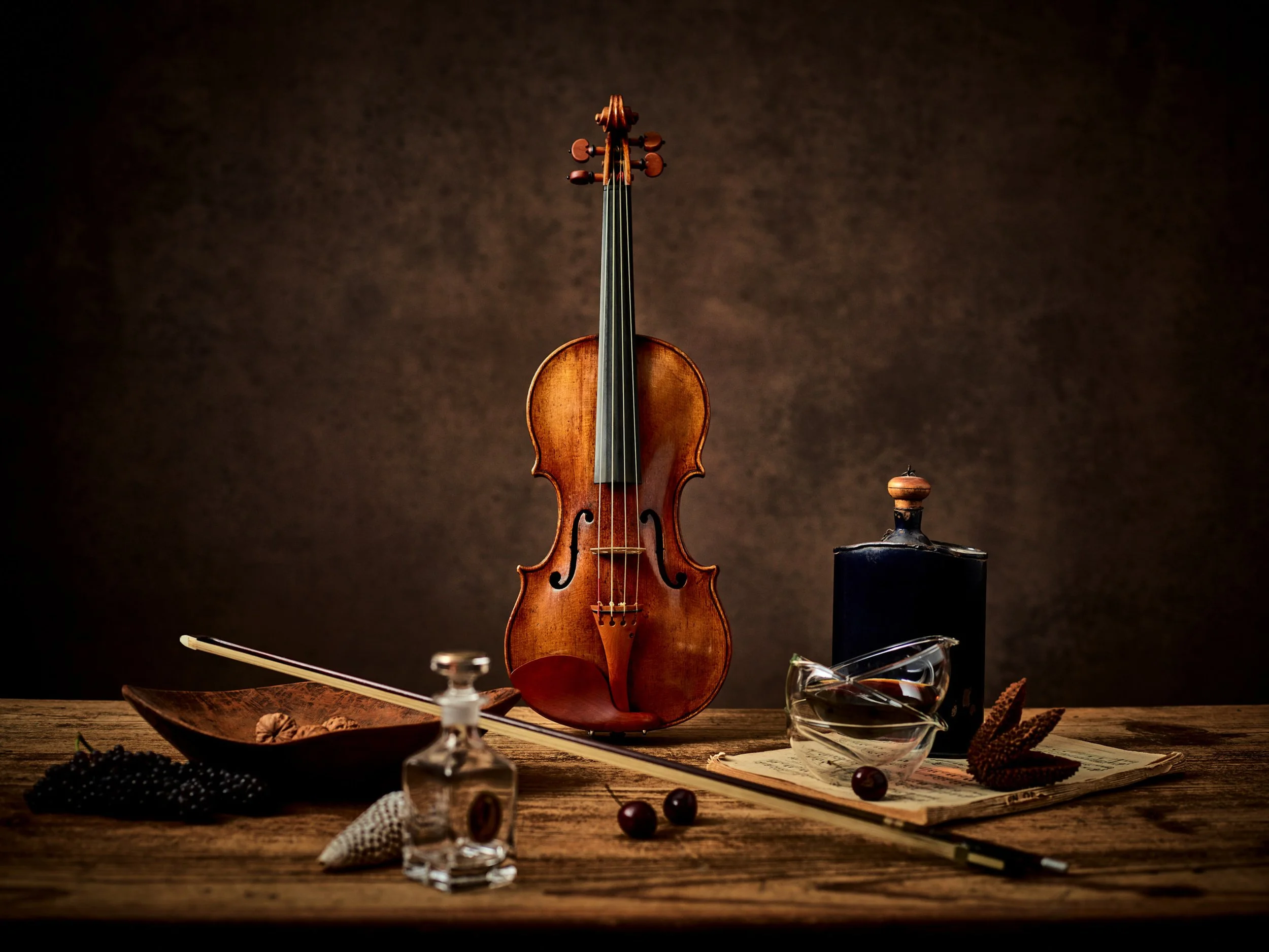 A solitary violin stands on a dark, textured background, surrounded by various objects on a wooden surface, including a bowl of walnuts, a small glass, a rolled piece of paper, and a dark bottle. The scene has a warm, vintage feel with dramatic light