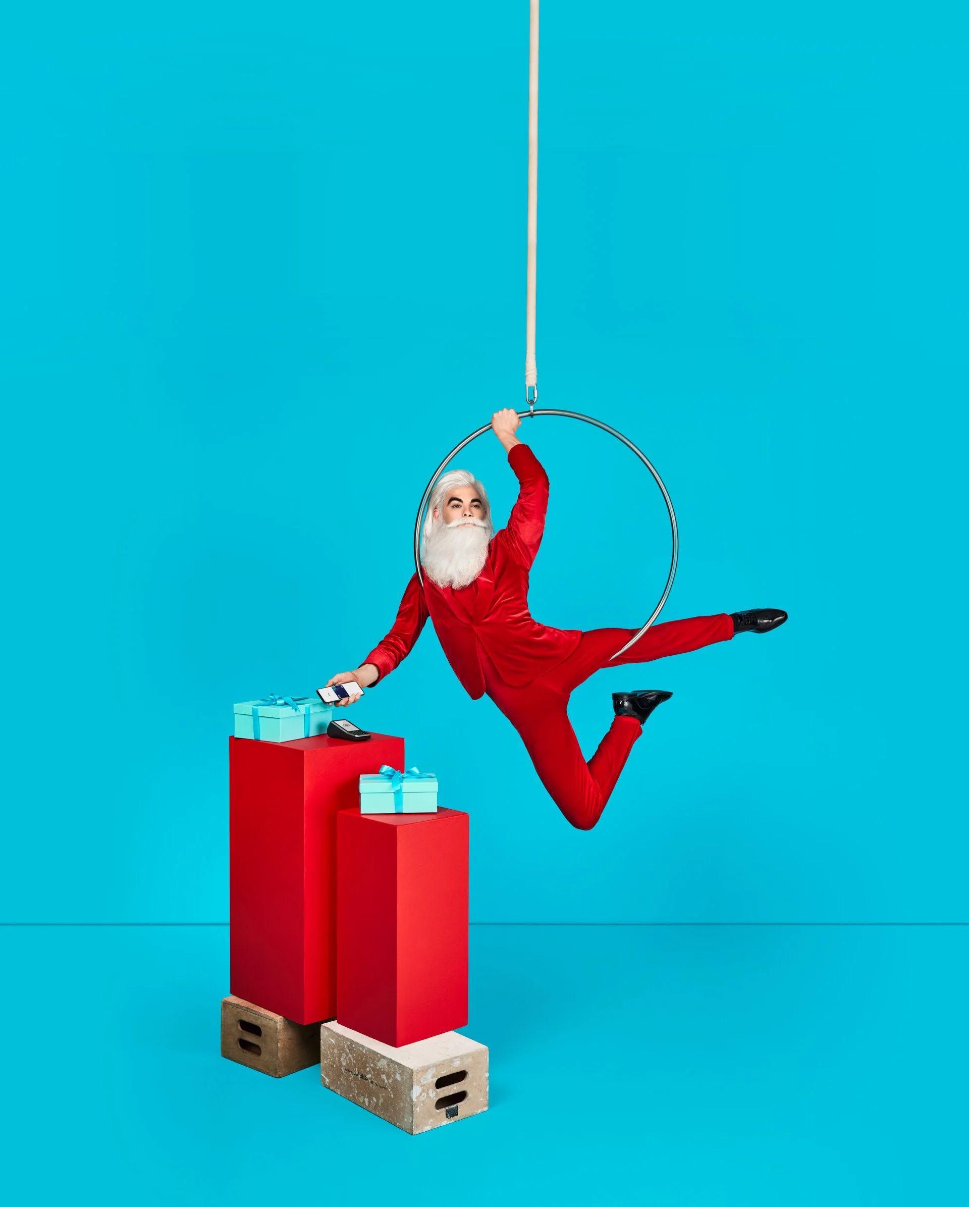 Santa Claus in a red suit doing a balancing pose on a gymnastic hoop hanging from the ceiling, with a series of colorful gift boxes and a red pedestal on his right, in front of a blue background.