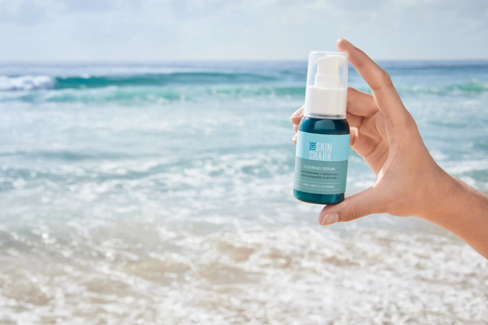 Hand holding a bottle of skincare serum labeled 'The Skin Shark' with the beach and ocean in the background.