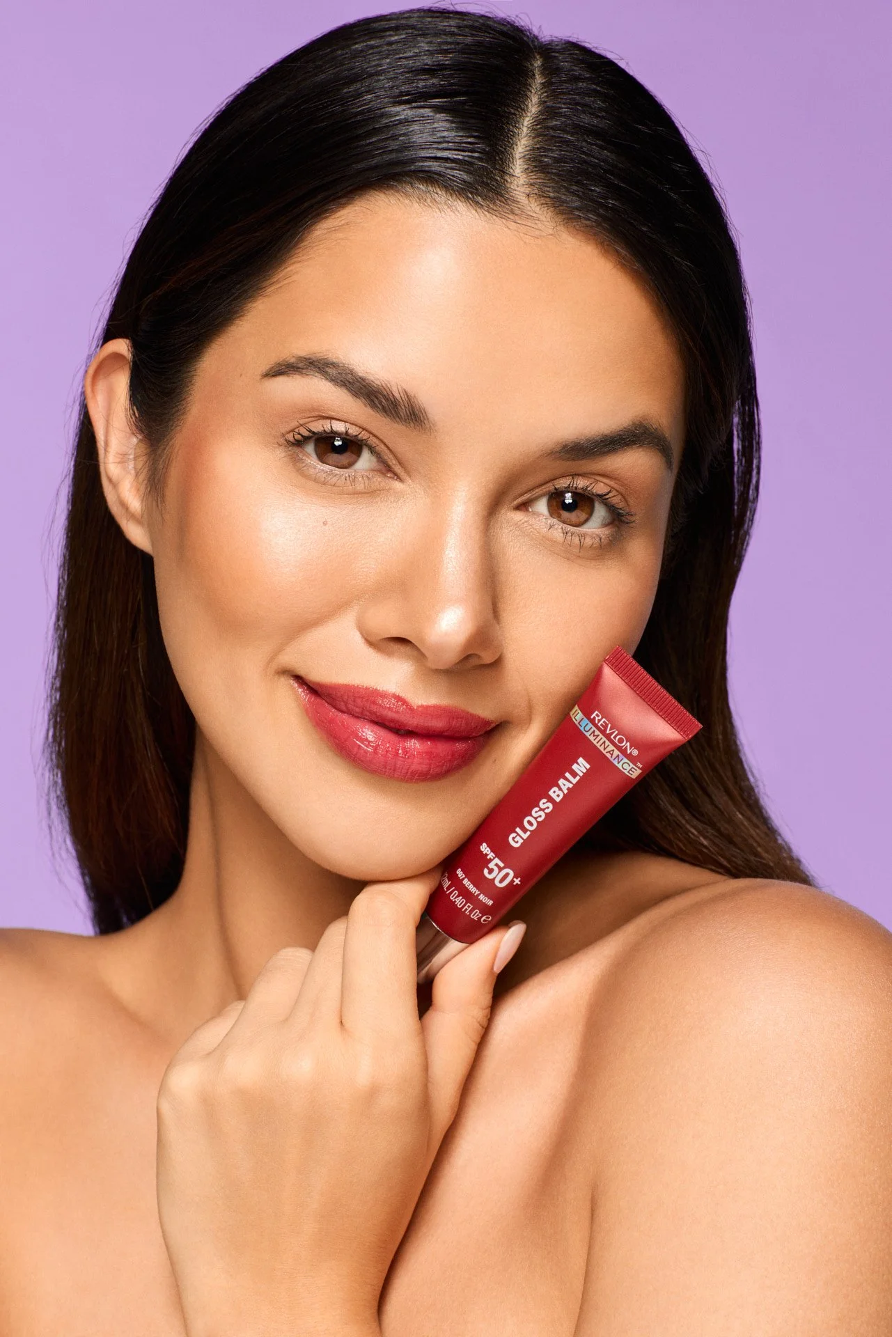 A woman with smooth skin, brown hair, and makeup holding a Revlon lip balm tube labeled SPF 50+ close to her face, smiling softly against a purple background.