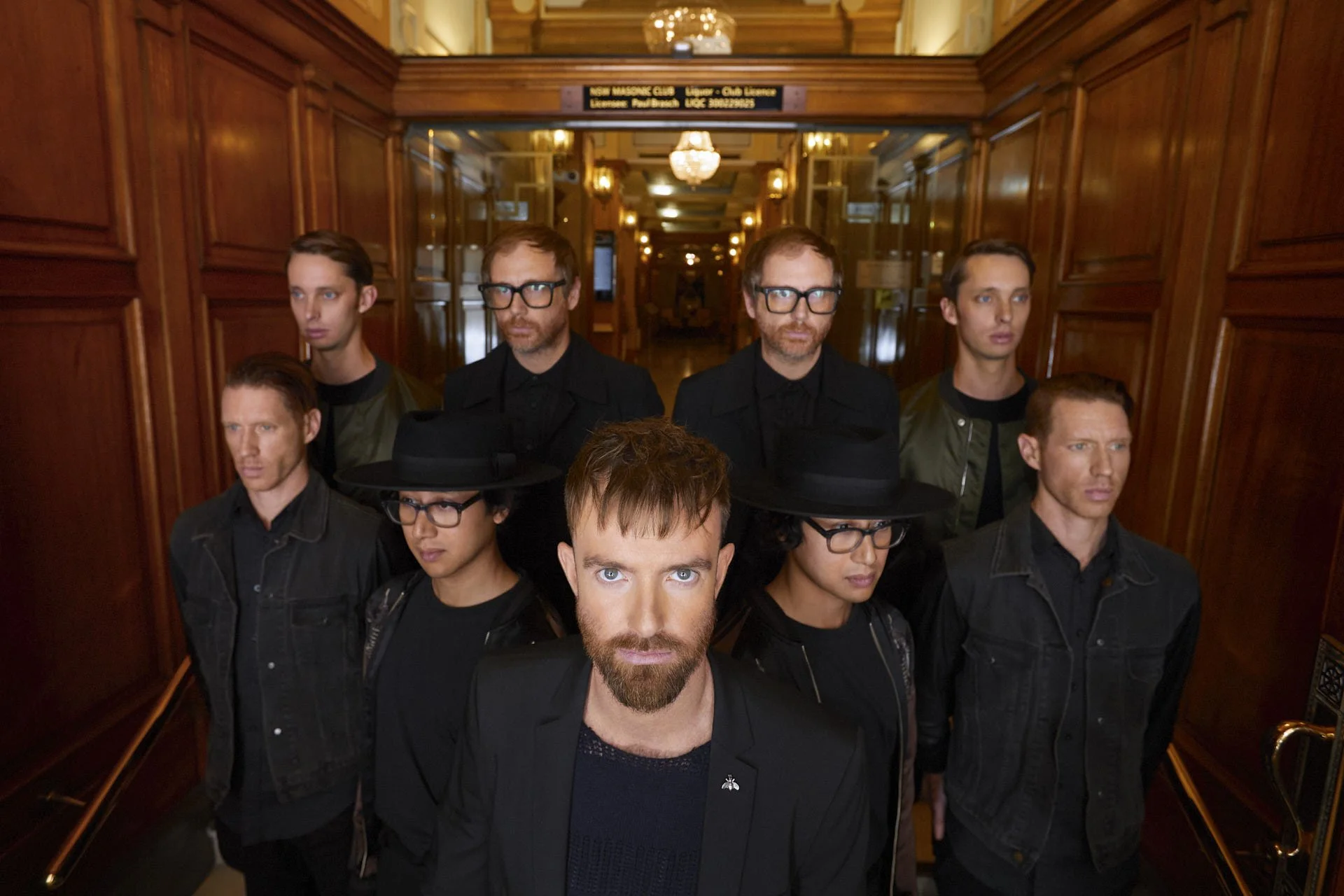 Group of ten men standing in an elevator with wood-paneled walls. The man in front has a beard and piercing blue eyes, while others wear glasses and dark clothing. The background shows a chandelier and a sign above the elevator door.