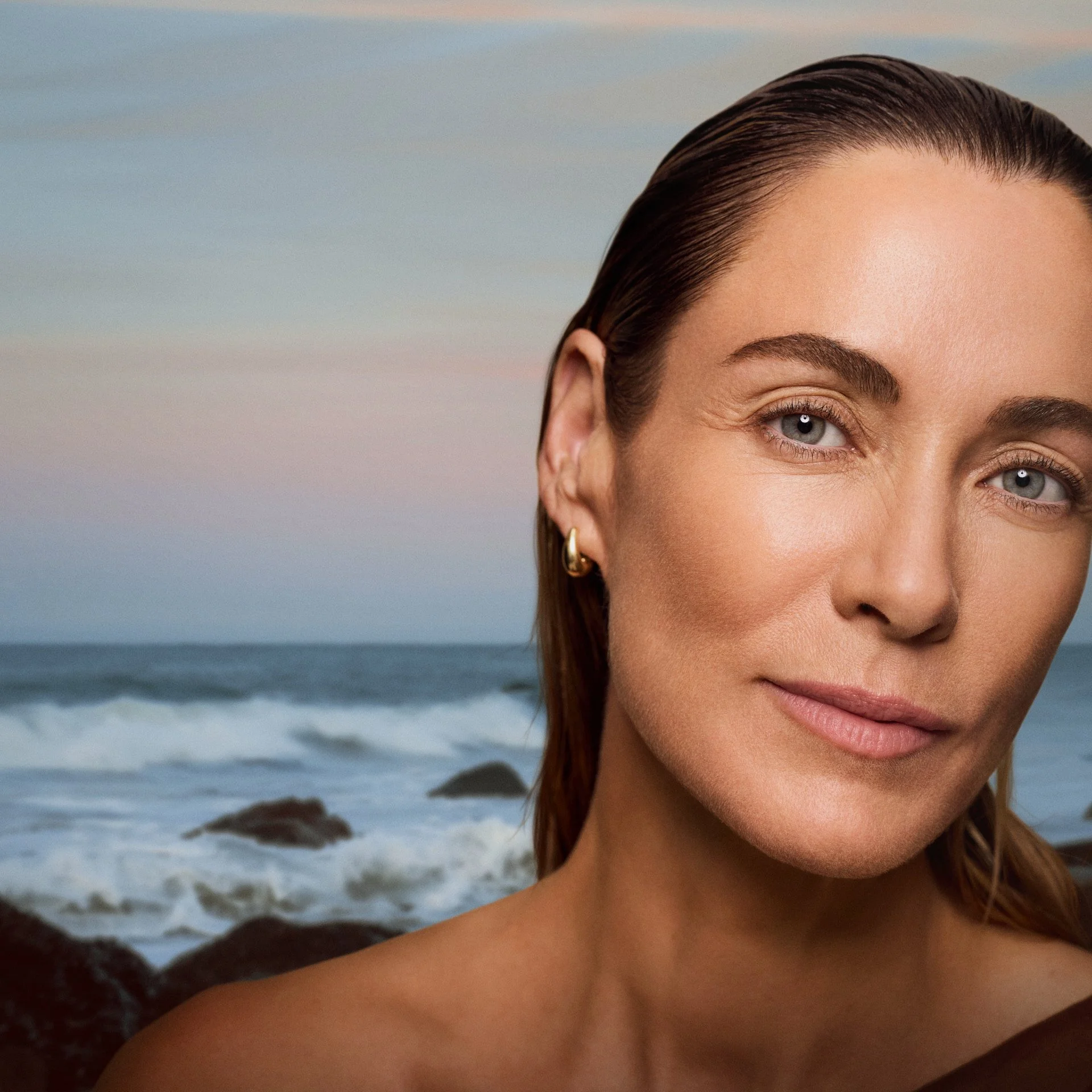Close-up of a woman's face, showing her eye, eyebrow, and part of her nose, with a beach and ocean in the background.