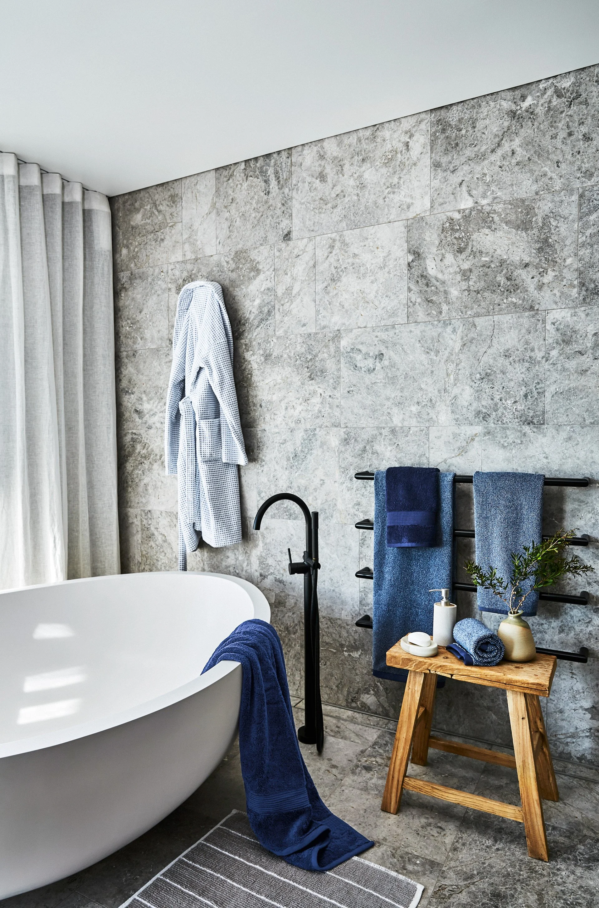 Modern bathroom with a white bathtub, gray stone walls and floor, and blue and white towels hanging on a black towel rack. A wooden table with toiletries and decor is next to the tub, and a gray striped rug on the floor.
