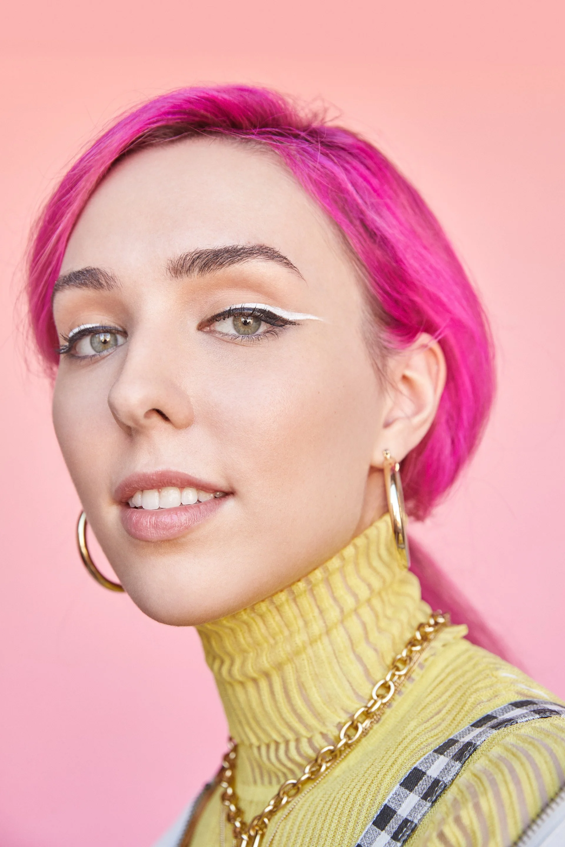 Close-up of a woman with short pink hair and green eyes, wearing yellow turtleneck and gold jewelry, against a pink background.