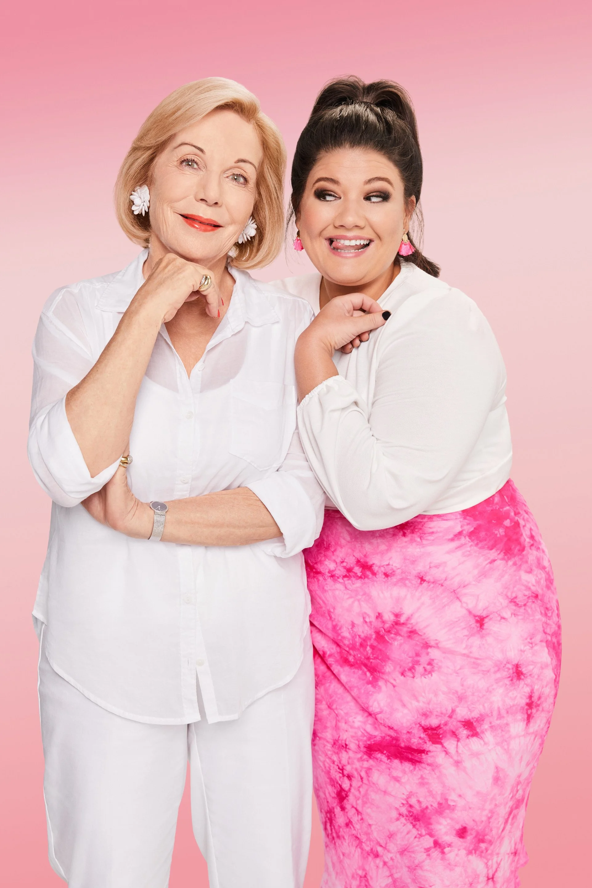 Two women, one older with blonde hair and one younger with dark hair, smiling and standing close together against a pink background. The older woman wears a white shirt with white floral earrings and red lipstick, while the younger woman wears a whit